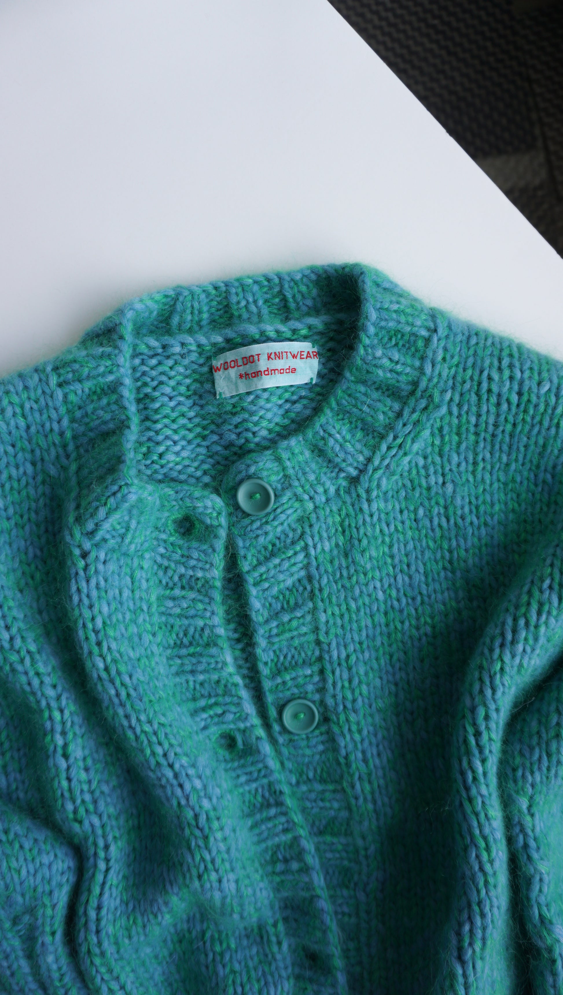Close-up of a teal knitted garment with a brand label on a white surface