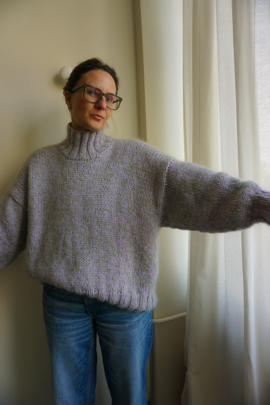 Person wearing a light purple sweater and blue jeans standing in front of a white curtain.