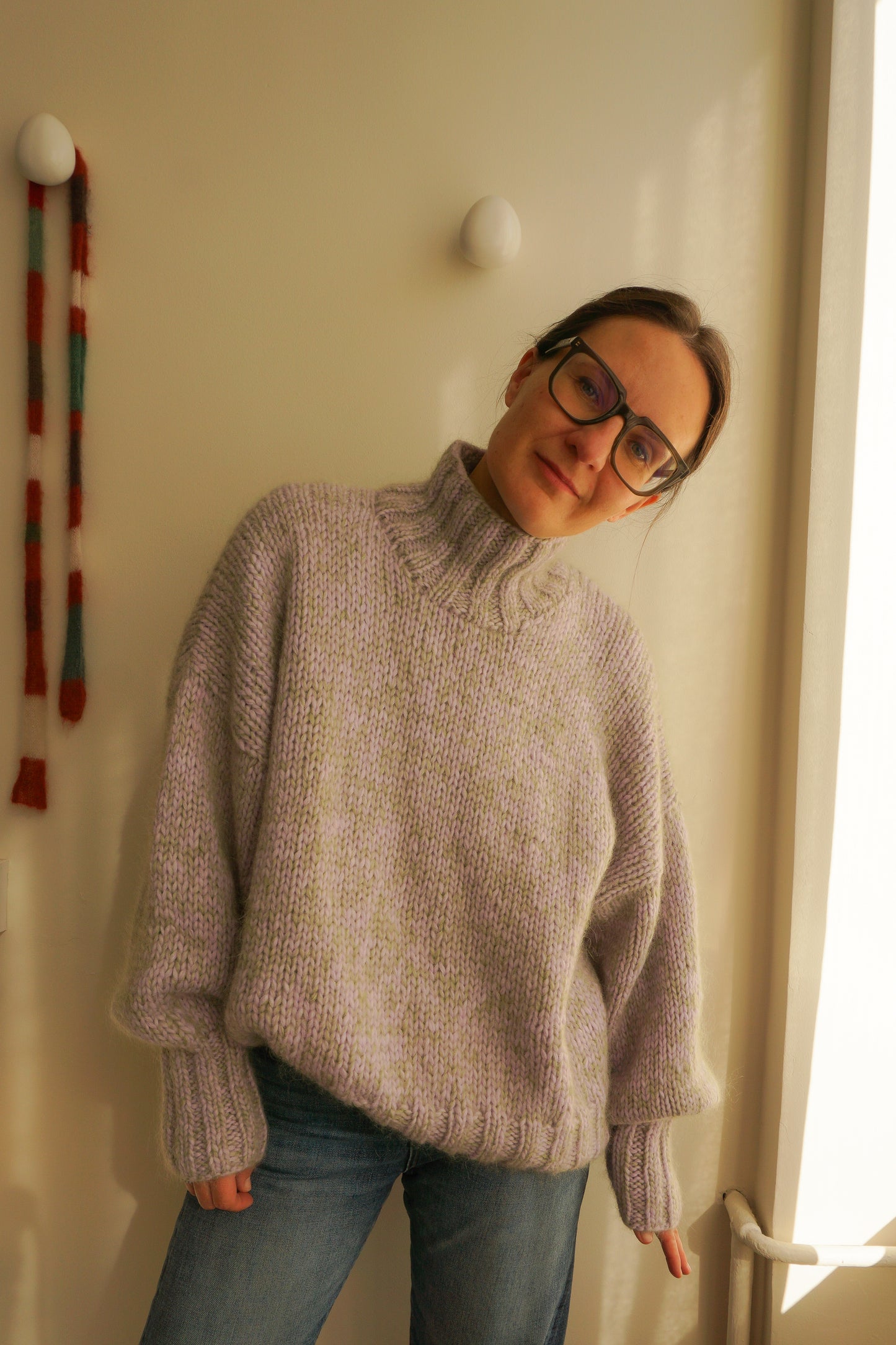 Person wearing a light purple sweater and glasses standing in a room with a light-colored wall and a window.