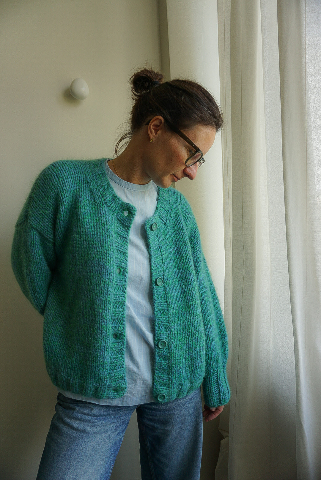 Woman wearing a teal knitted cardigan and glasses, standing in a room with white curtains.