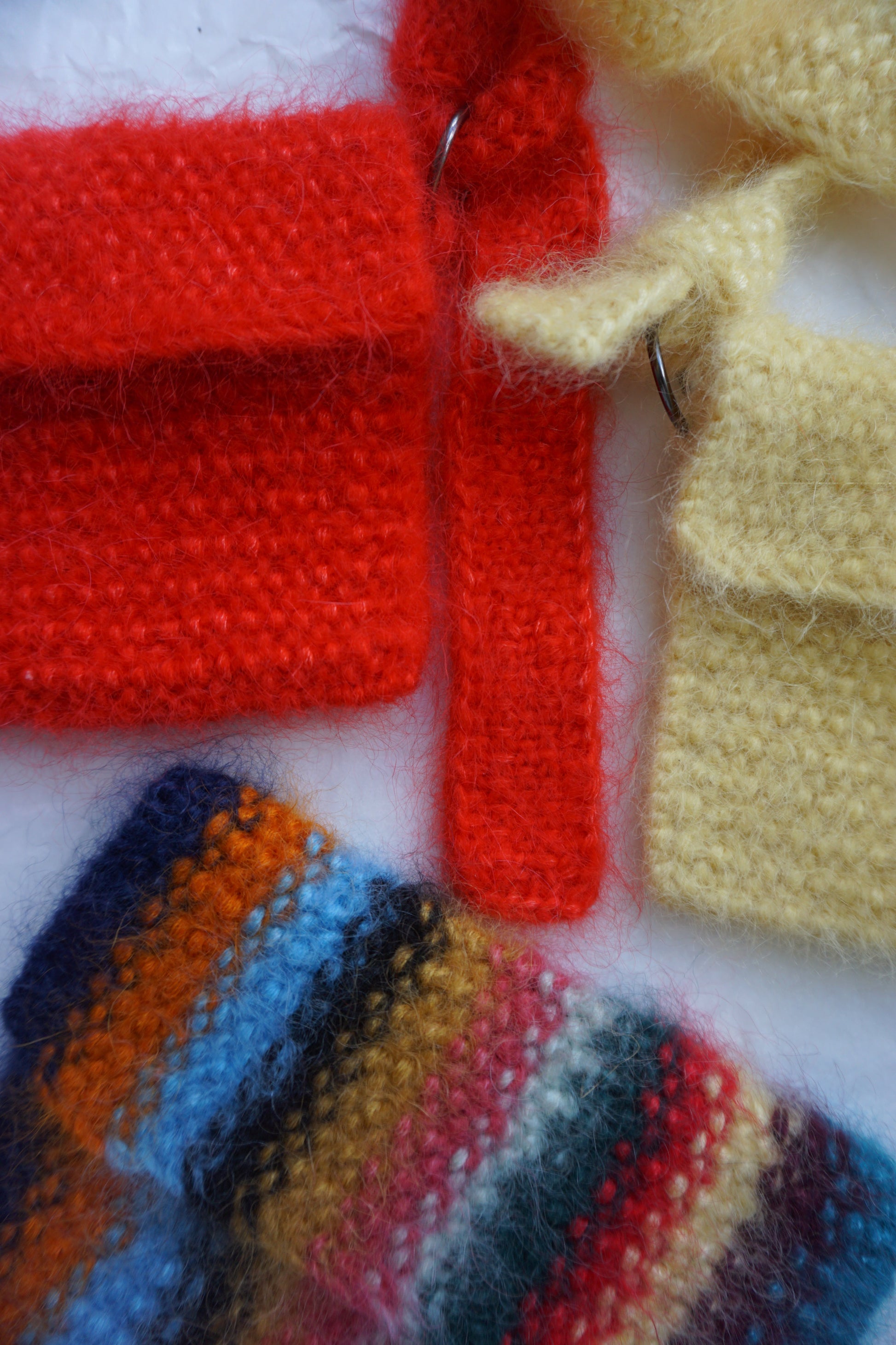 Close-up of knitted textile pieces in several colors, including pale yellow, bright red, and a striped multicolor section with blue, orange, yellow, red, and dark navy tones, laid on a white background.