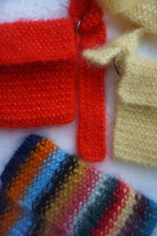 Close-up of knitted textile pieces in several colors, including pale yellow, bright red, and a striped multicolor section with blue, orange, yellow, red, and dark navy tones, laid on a white background.