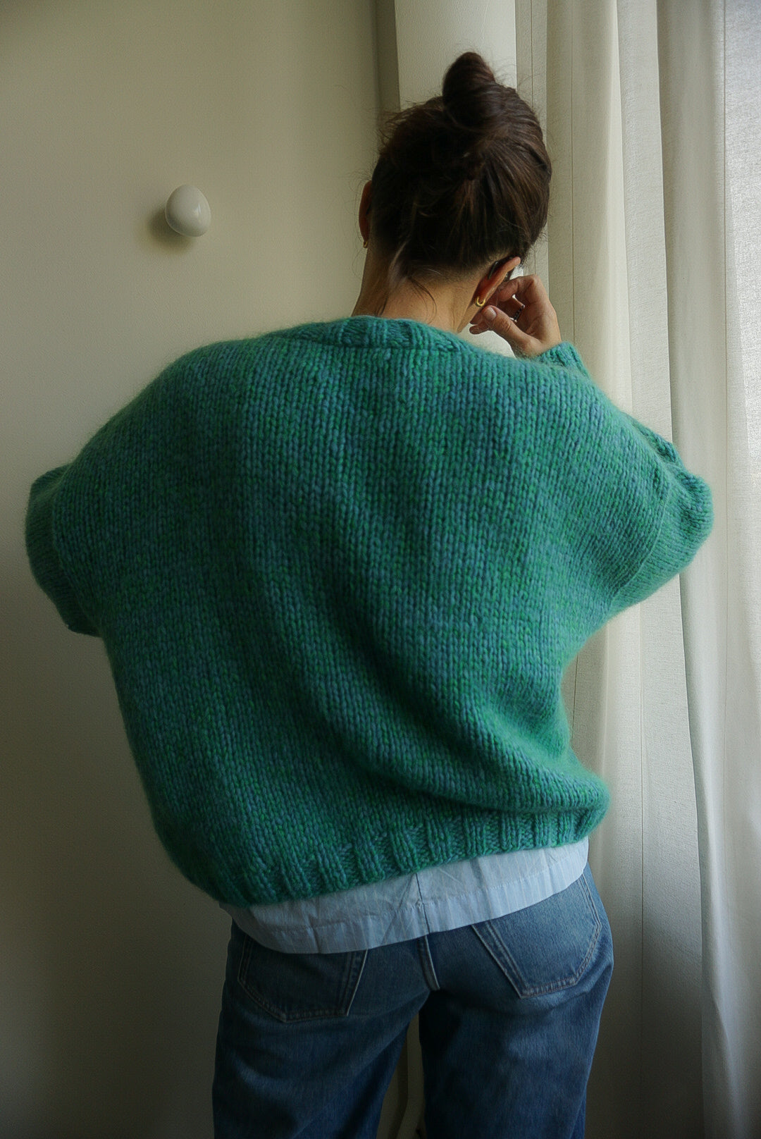 Person wearing a teal sweater and blue jeans standing in front of a white curtain.
