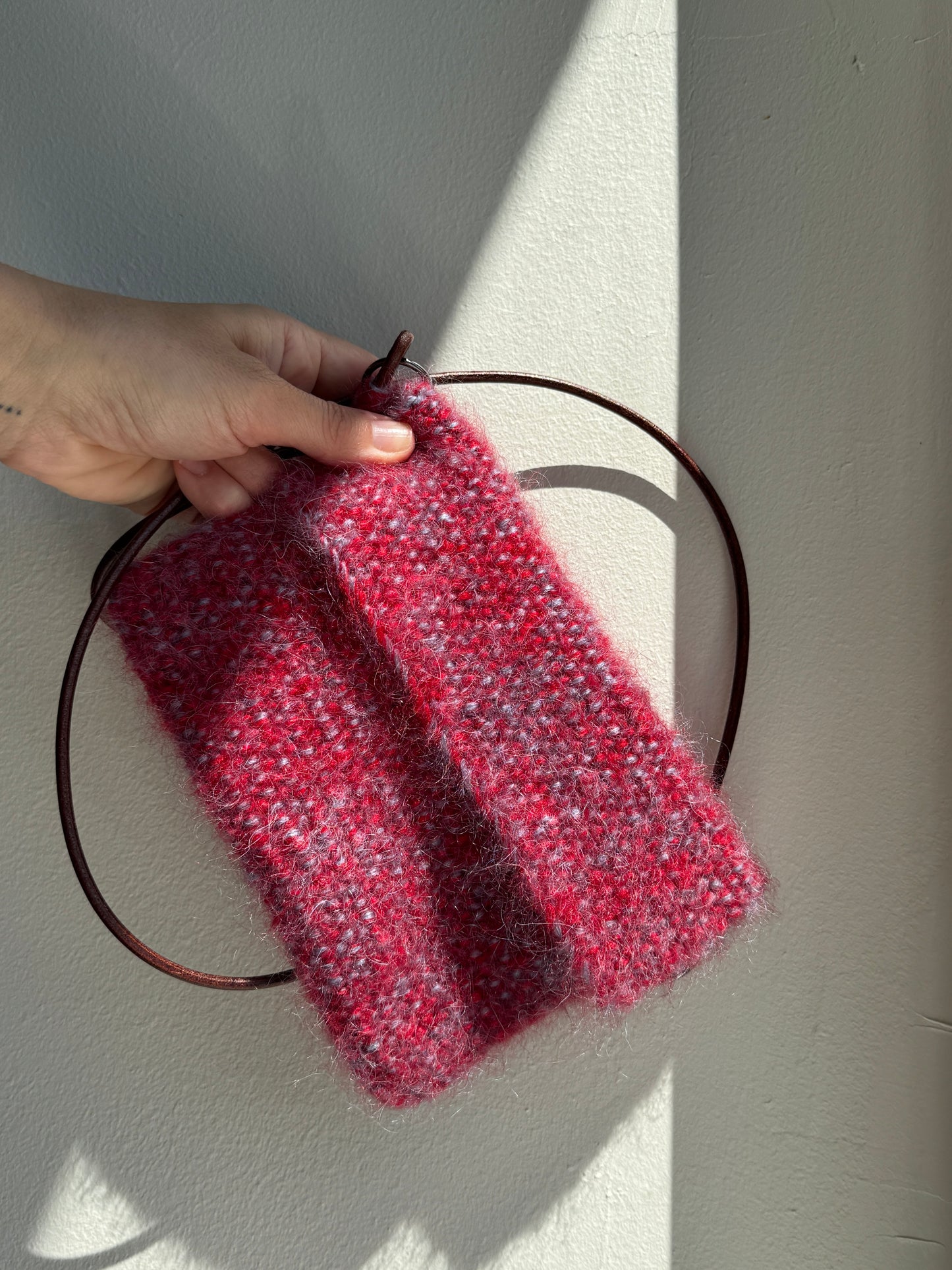 A hand holding a small chunky knit  bag in a heathered red and pink colorway, draped over a thin dark brown circular ring bag against a sunlit off-white wall.