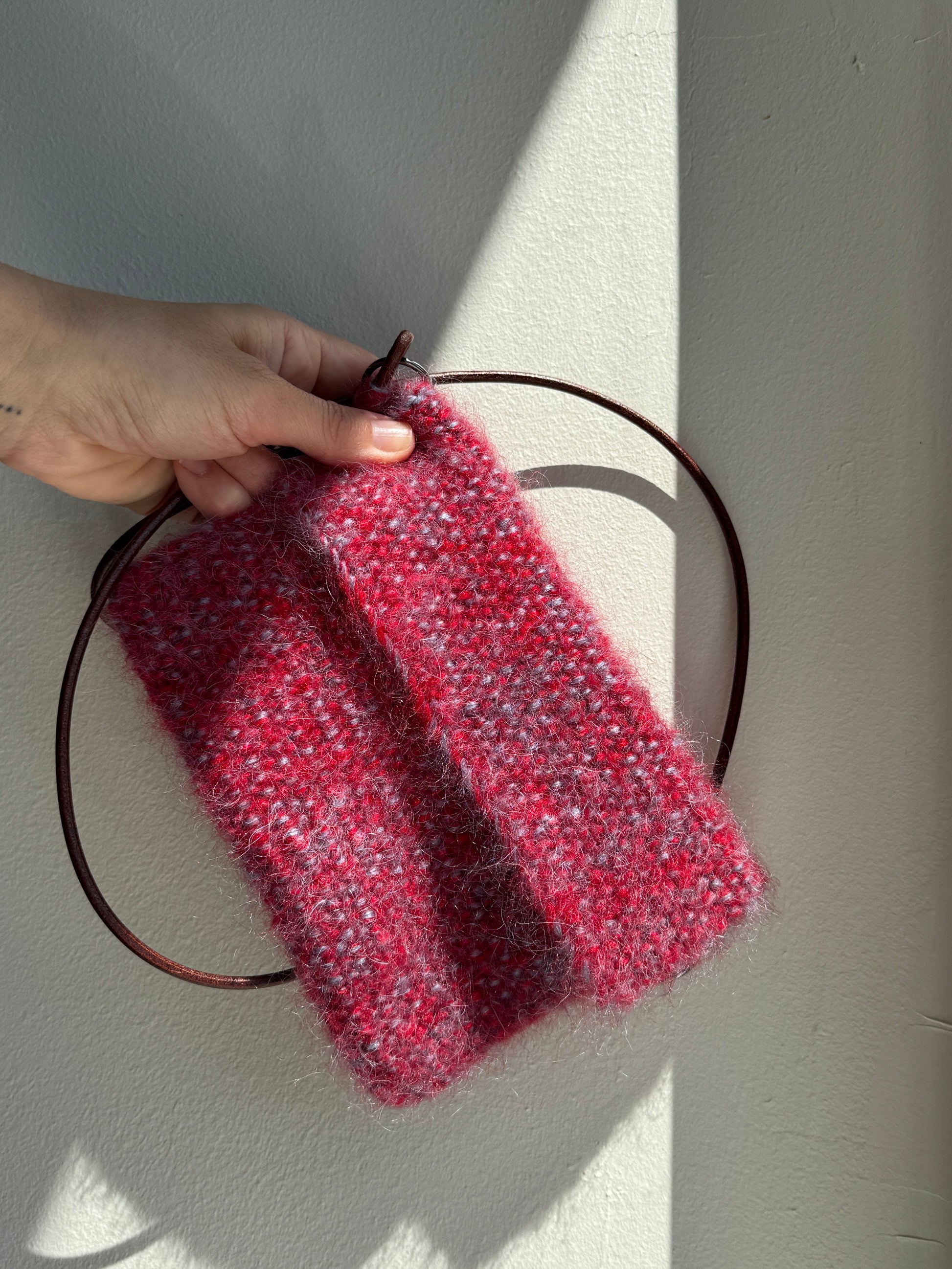 A hand holding a small chunky knit  bag in a heathered red and pink colorway, draped over a thin dark brown circular ring bag against a sunlit off-white wall.