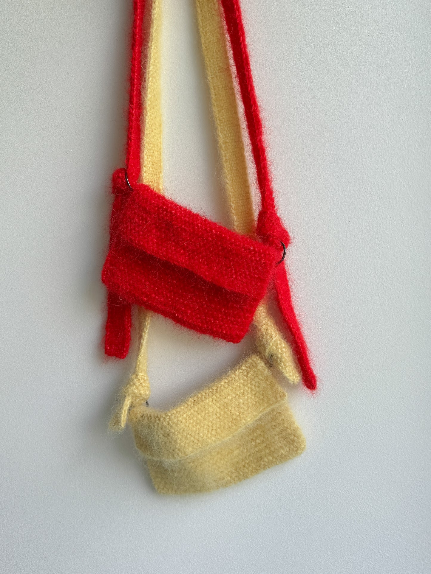 A small handmade crossbody bag made from textured yarn in red and pale yellow, hanging against a plain light gray background.