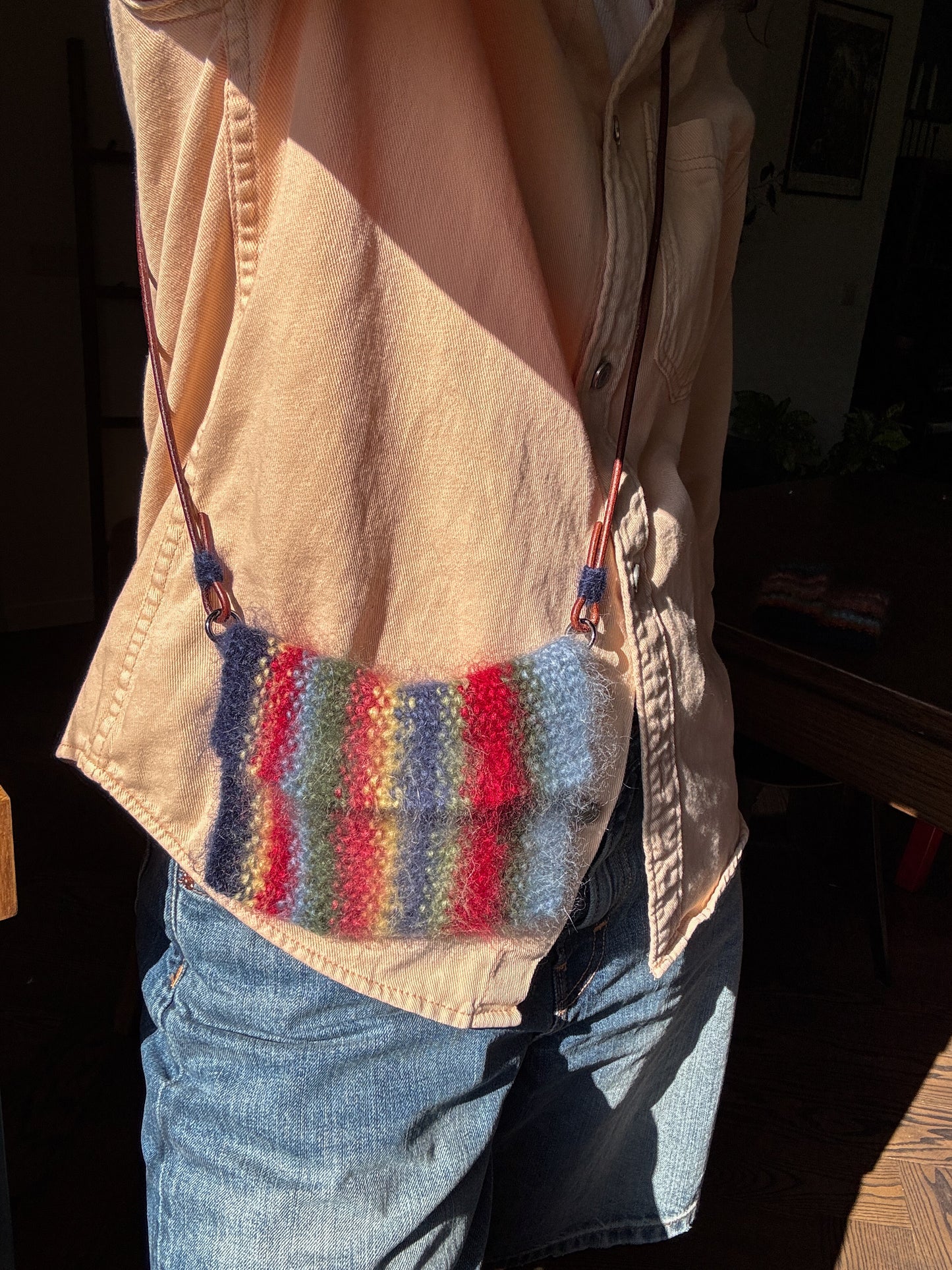 A person wearing a beige button-up shirt and blue jeans carries a small knitted shoulder bag with vertical stripes in blue, green, red, and pink. The photo is taken in warm, direct sunlight with a dark, shadowed indoor background.