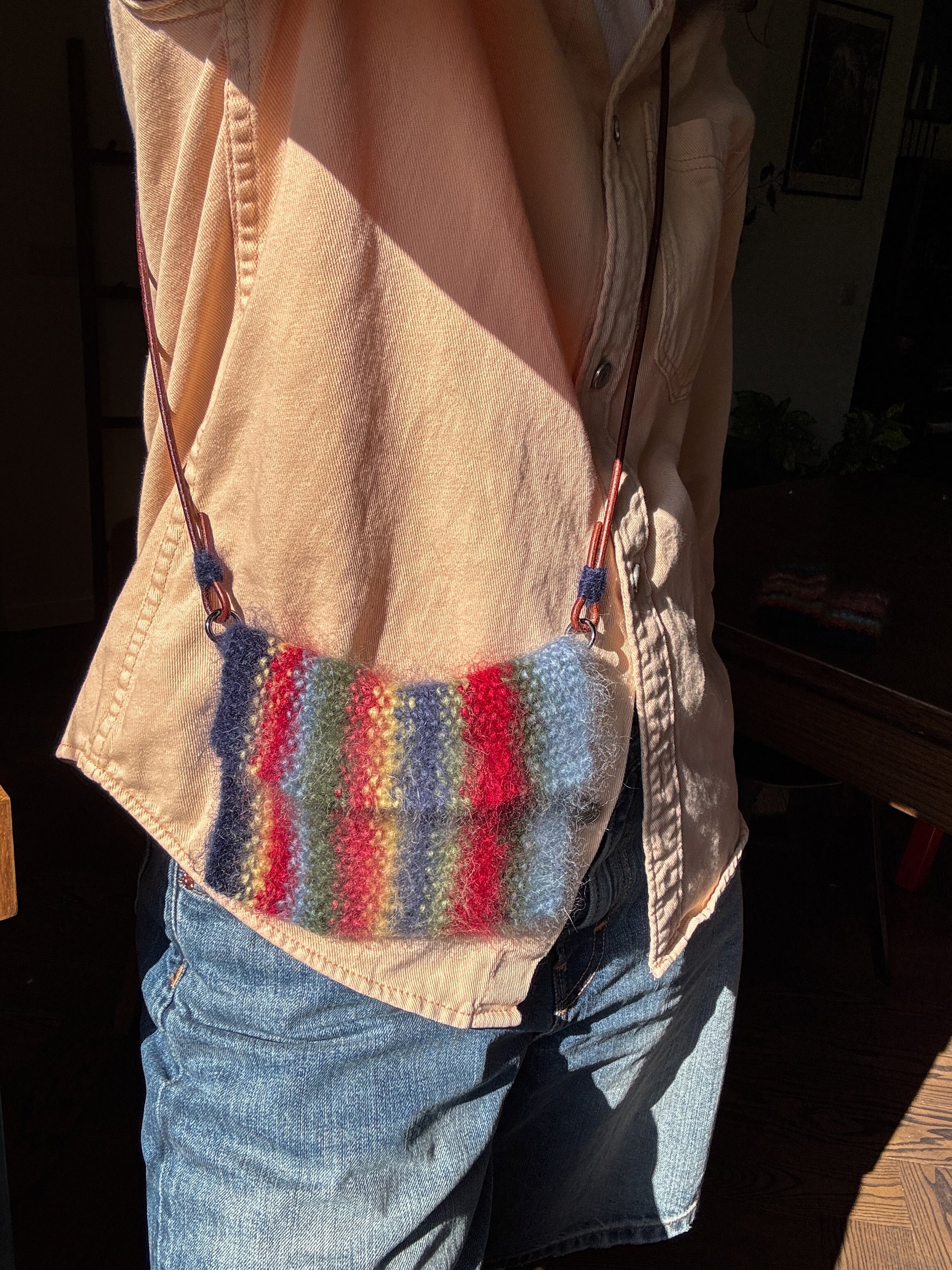 A person wearing a beige button-up shirt and blue jeans carries a small knitted shoulder bag with vertical stripes in blue, green, red, and pink. The photo is taken in warm, direct sunlight with a dark, shadowed indoor background.