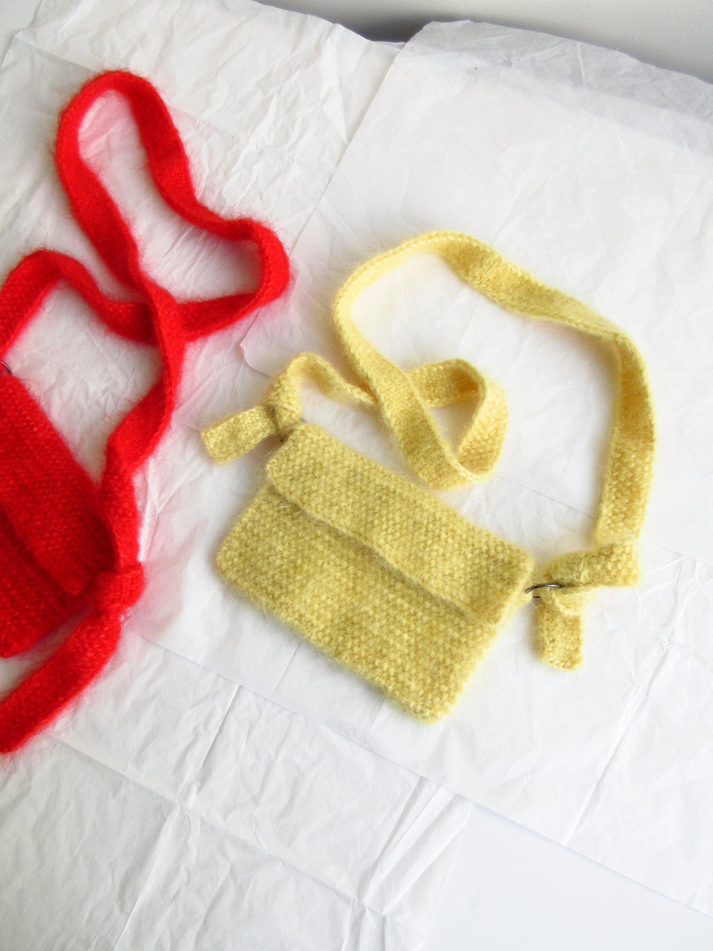 A small knitted yellow crossbody bag with a matching strap, placed on a white textured surface alongside a red knitted strap or accessory.